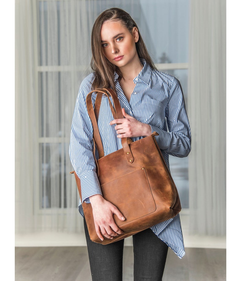 May include: A brown leather tote bag with a large front pocket and a zipper closure. The bag has a long strap that can be worn over the shoulder.
