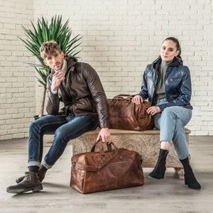 May include: Two people are sitting on a wooden bench with two brown leather travel bags. The person on the left is wearing a brown leather jacket and blue jeans. The person on the right is wearing a blue denim jacket and blue jeans.