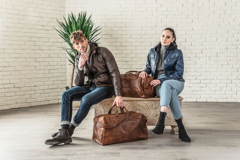 May include: Two people are sitting on a wooden bench with two brown leather travel bags. The person on the left is wearing a brown leather jacket and blue jeans. The person on the right is wearing a blue denim jacket and blue jeans.