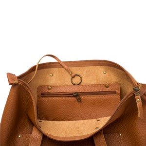 May include: A brown leather tote bag with a zipper closure and a small zippered pocket inside. The bag has a metal ring attached to the inside.