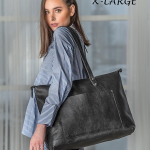 May include: A black leather tote bag with a zipper closure and a front pocket. The bag is being held by a person wearing a blue and white striped shirt and black pants. The text "BLACK X-LARGE" is visible in the top right corner of the image.