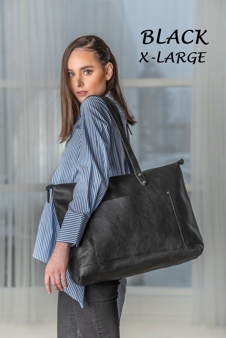 May include: A black leather tote bag with a zipper closure and a front pocket. The bag is being held by a person wearing a blue and white striped shirt and black pants. The text "BLACK X-LARGE" is visible in the top right corner of the image.