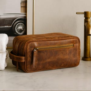 May include: A brown leather toiletry bag with a zipper closure and a handle. The bag is rectangular and has a distressed finish. The bag is sitting on a white marble surface.