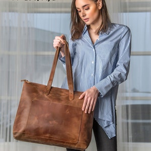 May include: A brown leather tote bag with a zipper closure and a front pocket. The bag is labeled "SADDLE X-LARGE".