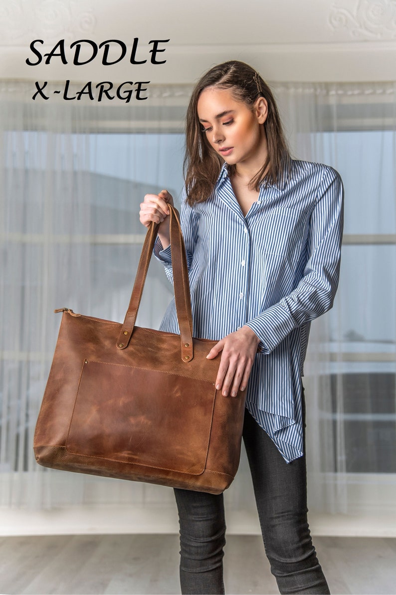 May include: A brown leather tote bag with a zipper closure and a front pocket. The bag is labeled "SADDLE X-LARGE".