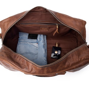 May include: A brown leather duffel bag with a zippered closure. The bag is open and contains a pair of blue jeans and a bottle of perfume.