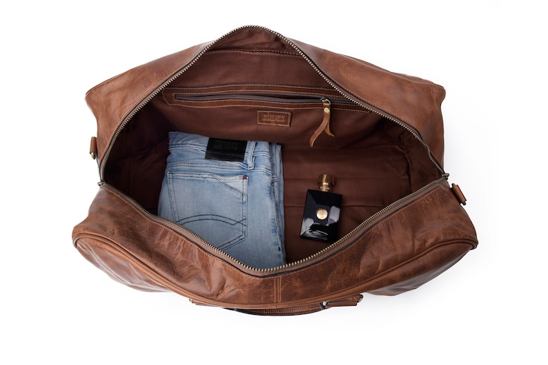 May include: A brown leather duffel bag with a zippered closure. The bag is open and contains a pair of blue jeans and a bottle of perfume.