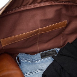May include: The inside of a brown leather duffel bag with a zipper closure. The bag has a brown fabric lining and a brown leather pocket on the inside. The bag is filled with a pair of blue jeans, a black shirt, and a pair of brown leather shoes.