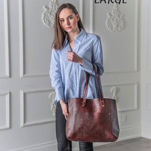May include: A woman wearing a blue and white striped shirt and black jeans is holding a large brown leather tote bag. The bag has a top zipper closure and two handles. The bag is labeled "TWO TONE OX RED LARGE".