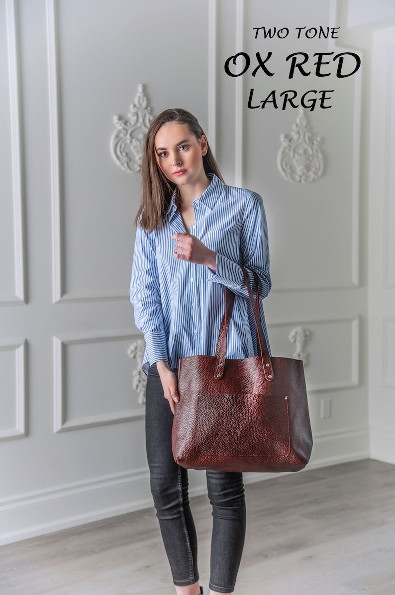 May include: A woman wearing a blue and white striped shirt and black jeans is holding a large brown leather tote bag. The bag has a top zipper closure and two handles. The bag is labeled "TWO TONE OX RED LARGE".
