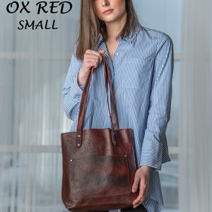May include: A woman is holding a brown leather tote bag with a blue and white striped shirt. The bag has a flap with a pocket and is made of textured leather. The text "TWO TONE OX RED SMALL" is visible in the top left corner of the image.