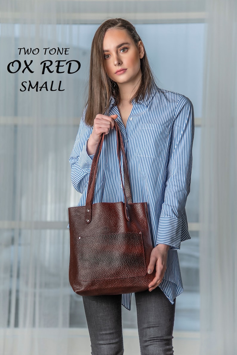 May include: A woman is holding a brown leather tote bag with a blue and white striped shirt. The bag has a flap with a pocket and is made of textured leather. The text "TWO TONE OX RED SMALL" is visible in the top left corner of the image.