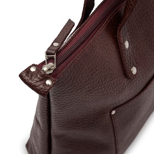 May include: A burgundy leather tote bag with a zipper closure and a small pocket on the front. The bag has silver hardware and a textured leather finish.