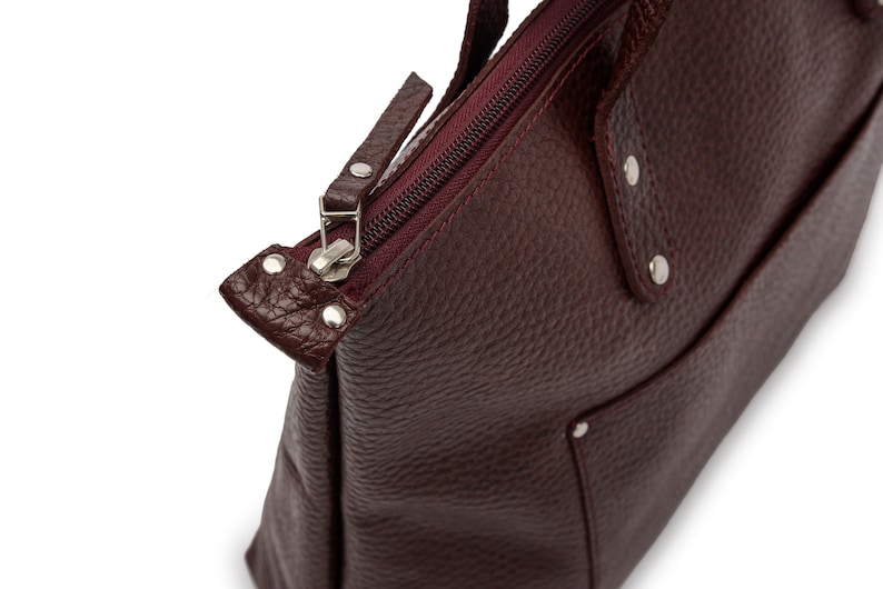 May include: A burgundy leather tote bag with a zipper closure and a small pocket on the front. The bag has silver hardware and a textured leather finish.