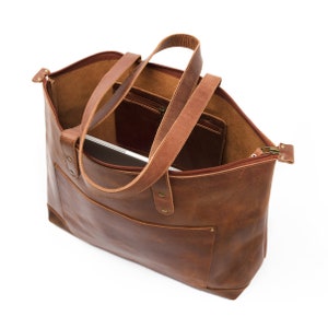 May include: A brown leather tote bag with a zipper closure and two top handles. The bag has a pocket on the inside and a laptop is visible inside the bag.