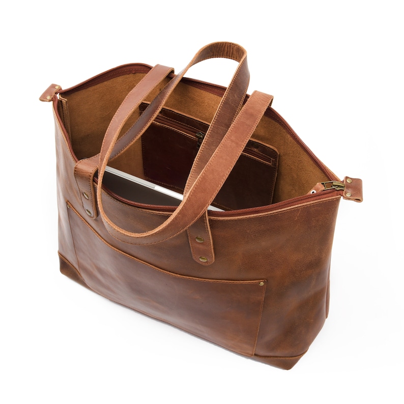 May include: A brown leather tote bag with a zipper closure and two top handles. The bag has a pocket on the inside and a laptop is visible inside the bag.