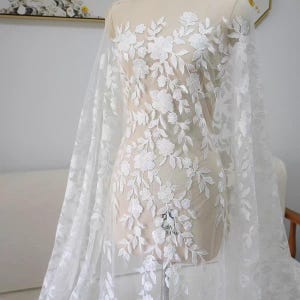 以下が含まれることがあります： マネキンに掛けられた白い花のレース生地。レースは、白い花と葉の繰り返し模様が特徴です。生地は透けており、マネキンが部分的に見えます。ブライダルやフォーマルウェアに最適です。