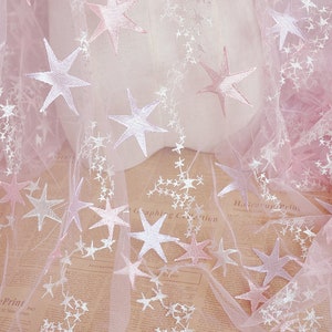 May include: Pink tulle fabric with white embroidered stars. The fabric has a subtle ombre effect, with the pink color fading to a lighter shade.