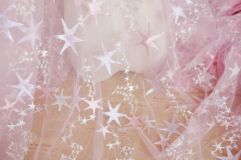 May include: Pink tulle fabric with white embroidered stars. The fabric has a subtle ombre effect, with the pink color fading to a lighter shade.