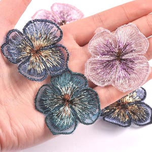 May include: Four floral embroidered patches featuring sheer mesh fabric with metallic thread detailing. The set includes two blue-toned flowers with gold centers, one light pink flower with a purple center, and one pale pink flower.