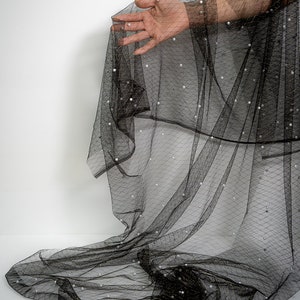 Puede incluir: Tela negra transparente con peque&ntilde;as perlas blancas. La tela est&aacute; drapeada sobre una mano.