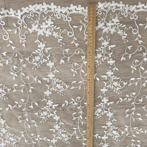 Puede incluir: Tela de encaje floral blanco con delicados patrones de flores y hojas. Una regla de madera muestra las medidas en centímetros. El encaje presenta un borde festoneado, adecuado para ropa de novia, manualidades o prendas.