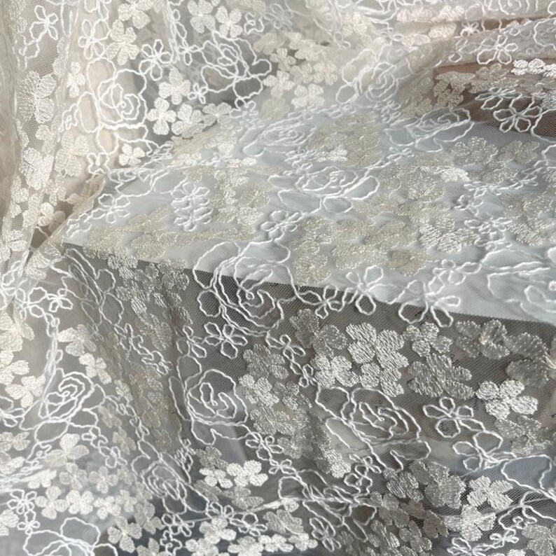 May include: White floral lace fabric with a delicate pattern of flowers and leaves. The lace is sheer and has a soft, flowing texture.
