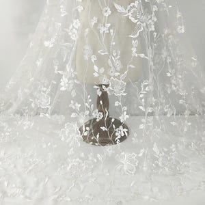 Puede incluir: Tela de encaje floral blanco con flores y vides bordadas. El delicado encaje está drapeado sobre un maniquí, mostrando el intrincado diseño. Perfecto para ropa de novia u otras ocasiones especiales.