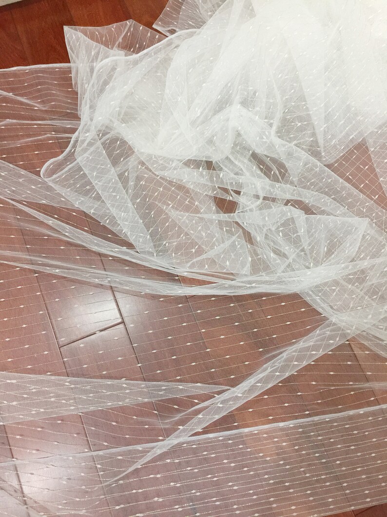 May include: White tulle fabric with a delicate pattern of small dots. The fabric is sheer and has a soft, flowing texture.