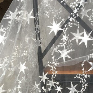 May include: White sheer fabric with a silver star pattern. The fabric is draped over a wooden surface.