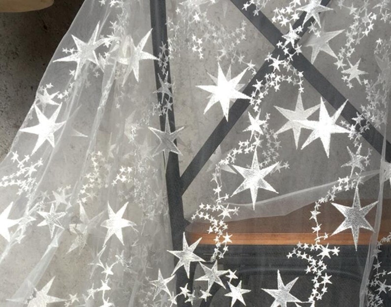 May include: White sheer fabric with a silver star pattern. The fabric is draped over a wooden surface.