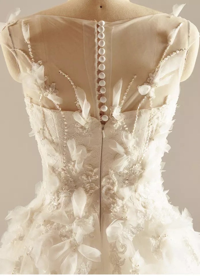May include: A white wedding dress with a sheer bodice and a skirt with layers of tulle and floral appliques. The back of the dress features a zipper closure and a row of buttons.
