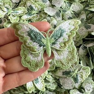 Könnte beinhalten: Nahaufnahme einer Hand, die eine grüne und weiße gestickte Schmetterlingsapplikation hält. Der Schmetterling hat ein detailliertes Design mit einer weißen Mitte und grünen Flügeln. Der Schmetterling ist von anderen ähnlichen Schmetterlingen umgeben.