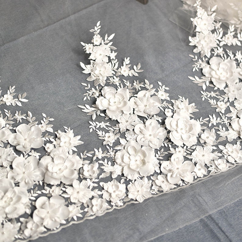 May include: White floral lace trim with 3D flowers and pearl accents. The lace is delicate and intricate, with a soft, flowing texture. The flowers are raised and have a realistic appearance. The pearls are small and scattered throughout the lace.