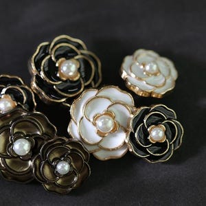 Può includere: Una collezione di gioielli a forma di fiore in nero, bianco e bronzo, con finiture dorate. Ogni fiore presenta un centro di perla finta. I gioielli sono esposti su uno sfondo scuro.