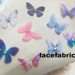 May include: Assortment of handmade fabric butterflies with rhinestones in various colors, including pink, blue, and purple. The butterflies are arranged on a white fabric background. The text "lacefabricArt" is visible in the lower right corner of the image.
