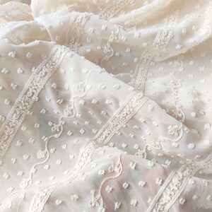 May include: White sheer fabric with a textured, dotted pattern and delicate embroidered lace trim. The fabric is soft and lightweight, perfect for creating garments or home decor.