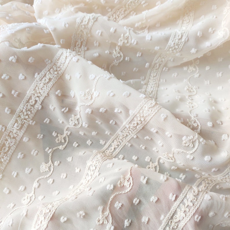 May include: White sheer fabric with a textured, dotted pattern and delicate embroidered lace trim. The fabric is soft and lightweight, perfect for creating garments or home decor.