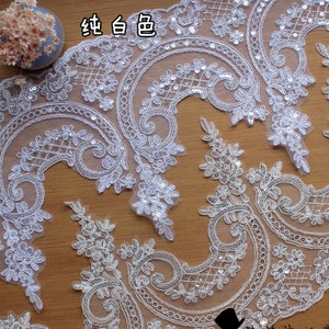 Puede incluir: Borde de encaje blanco con un delicado patrón floral y lentejuelas plateadas. El borde está sobre una superficie de madera.