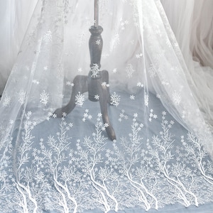 May include: White sheer fabric with an embroidered tree and snowflake design. The delicate pattern is visible against a light blue background. The fabric is draped over a wooden mannequin.