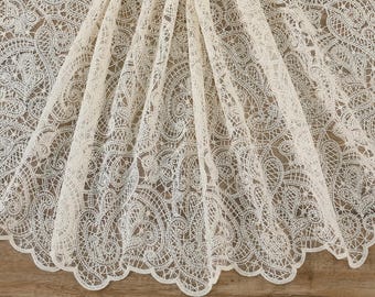 May include: Ivory-colored lace fabric with an intricate, openwork design. The fabric is draped, revealing the detailed patterns and scalloped edge. The lace features floral and geometric motifs, suitable for various craft projects.