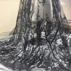 Peut inclure: Tissu en tulle noir avec un motif d'arbres brodés. Le tissu présente un motif répété d'arbres avec des détails complexes et de petits sequins éparpillés. Le tulle est drapé sur une forme, mettant en valeur sa texture et son design.