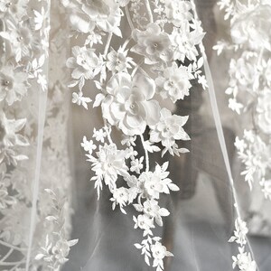 May include: White floral lace fabric with 3D flowers and pearl accents. The fabric is sheer and delicate, with a soft, romantic feel.