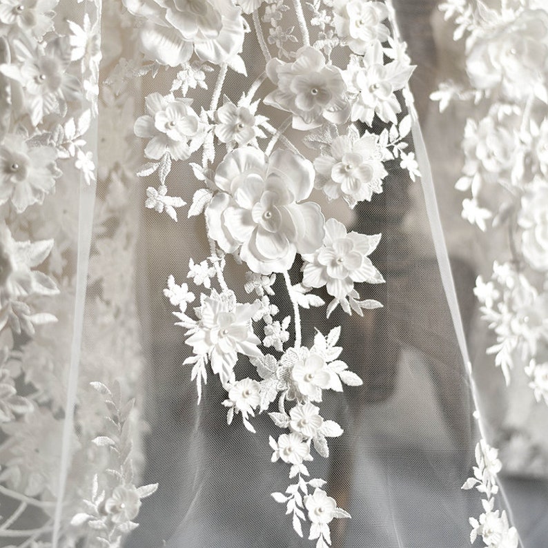 May include: White floral lace fabric with 3D flowers and pearl accents. The fabric is sheer and delicate, with a soft, romantic feel.