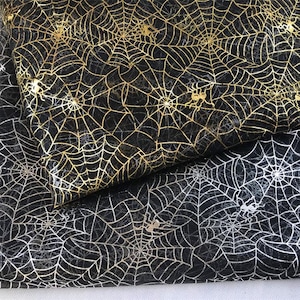 Puede incluir: Primer plano de tela negra con un diseño de telaraña dorada metálica. La tela tiene un acabado brillante y está doblada, revelando los intrincados detalles del patrón. Pequeñas siluetas de arañas están dispersas por el diseño de la telaraña. La tela es adecuada para manualidades temáticas de Halloween.