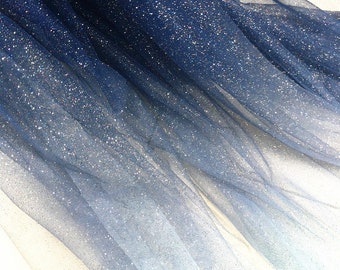 May include: Blue and white ombre tulle fabric with glitter. The fabric is sheer and has a soft, flowing texture.