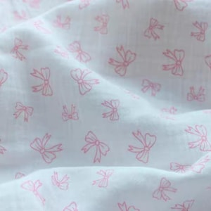 Könnte beinhalten: Weißer Stoff mit einem sich wiederholenden Muster aus rosa Schleifen. Die Schleifen sind rosa umrandet und über dem weißen Hintergrund verstreut. Der Stoff wirkt weich und leicht zerknittert, was auf ein leichtes Material hindeutet.