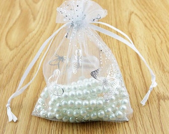 Puede incluir: Una bolsa de regalo de organza blanca transparente con purpurina plateada y diseños de mariposas. La bolsa está atada con una cinta de satén blanca y contiene una cadena de perlas blancas.