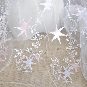May include: White tulle fabric with pink and white embroidered stars. The fabric is sheer and delicate, perfect for crafting or adding a touch of whimsy to any project.