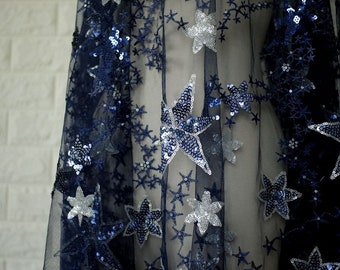 May include: Navy blue sheer fabric with silver and blue star-shaped sequins. The fabric is decorated with a repeating pattern of stars.